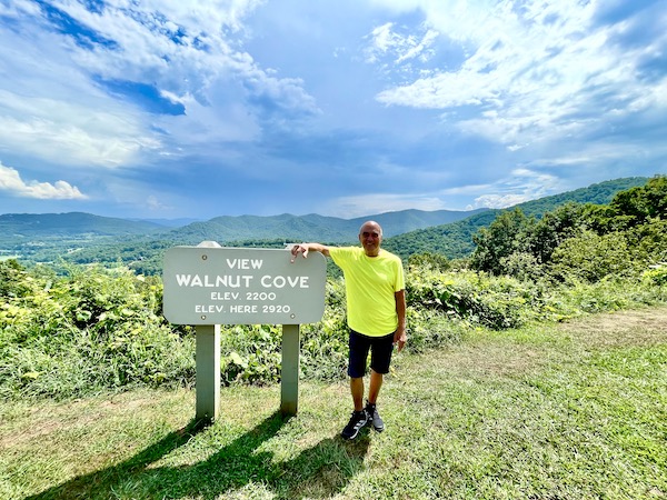 A man in a yellow T-shirt leaning on a gray wood sign with mountains and clouds behind him.