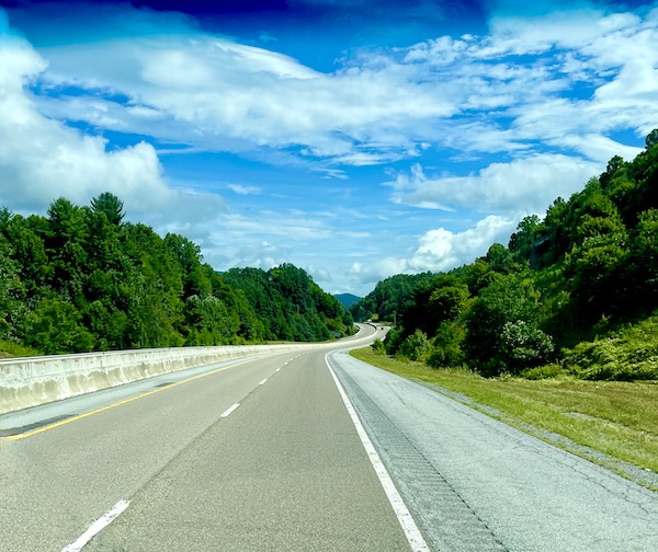 A curved highway lined with green trees and lots of blue sky with wispy white clouds.