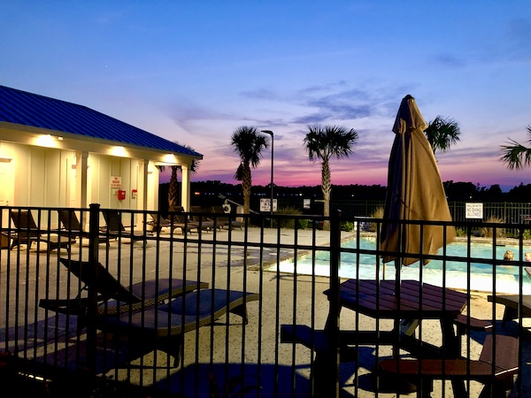 Sunset at a pool with palm trees and umbrella tables.
