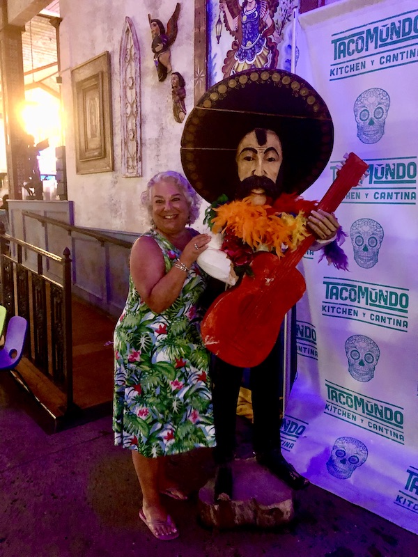 A silver haired woman in a tropical print dress next to a stature of a Mexican guitar player at Taco Mundo North Myrtle Beach