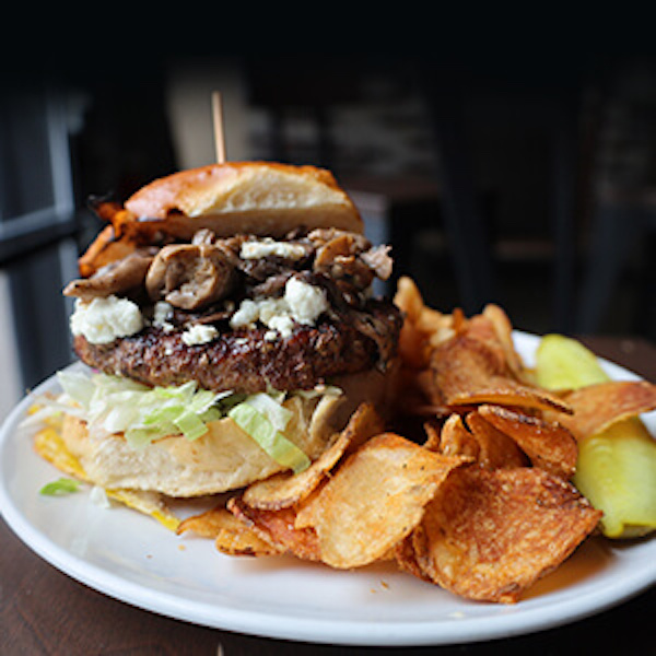 A round white plate topped with a huge burger with mushrooms and white cheese along with a side of fresh chips and pickle spear at Mudhen Brewing Wildwood NJ