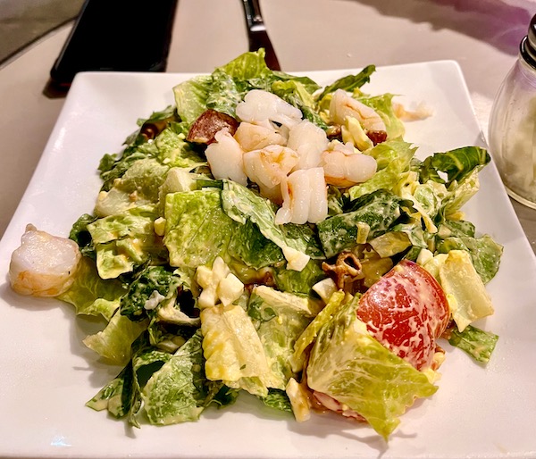 A square white plate topped with salad greens, tomato and shrimp with creamy dressing.