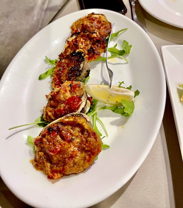 An oval white platter topped with 6 clamshells filled with clams and breadcrumbs and a few arugula leaves plus a wedge of lemon.