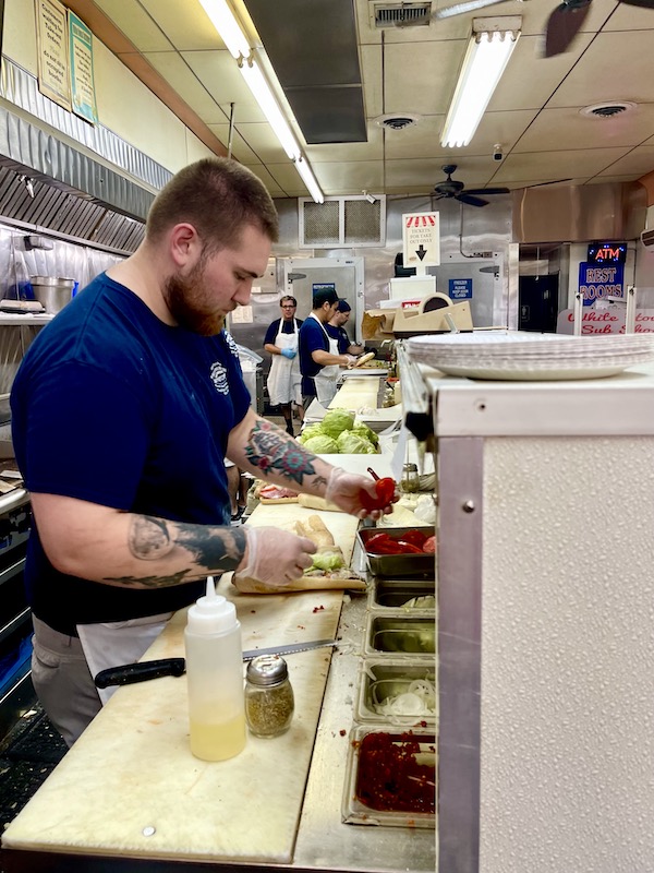 A counter where sub sandwiches are made and men standing behind it doing prep