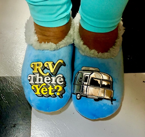A woman's feet with blue slippers. On one foot it says RV there yet? and on the other is an Airstream appliquéd.