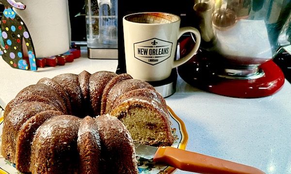 A glazed Sour Cream Coffee Cake sits on a yellow rimmed platter with an orange handled knife. In the background is a Keurig with a beige mug.