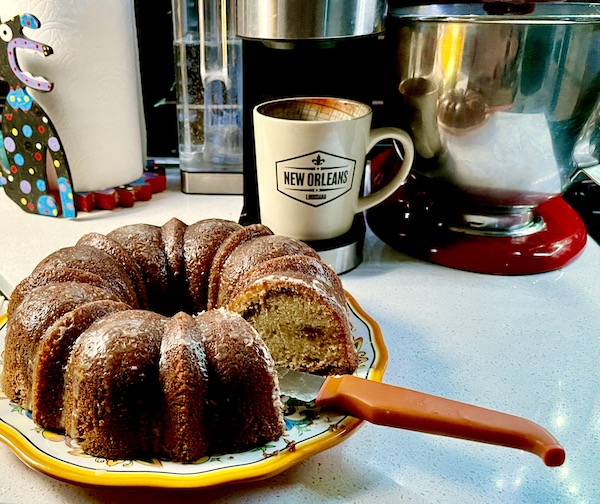 A glazed Sour Cream Coffee Cake sits on a yellow rimmed platter with an orange handled knife. In the background is a Keurig with a beige mug.