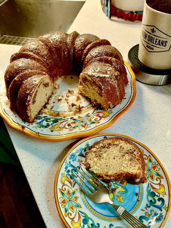 A Sour Cream Coffee Cake on a platter with a portion cut out. A single slice sits on a blue and yellow plate with a dessert fork.