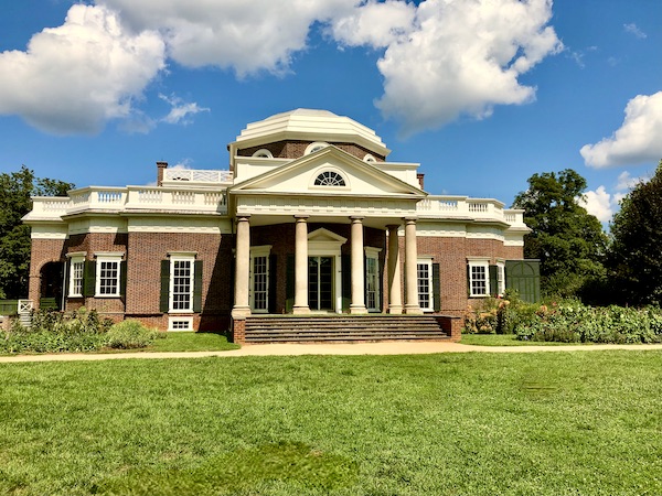 The rear of a red brick mansion with white pillars and lawn.