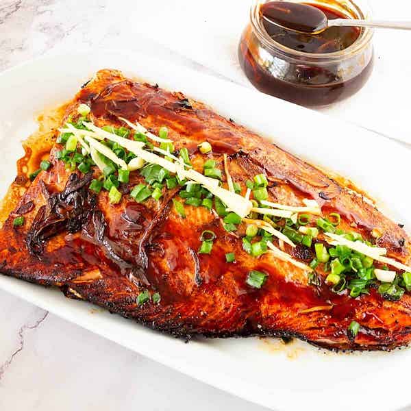 Baked teriyaki salmon on a white platter topped with greens and a dish of brown sauce, one of 7 Creative Ways to Serve Up Salmon