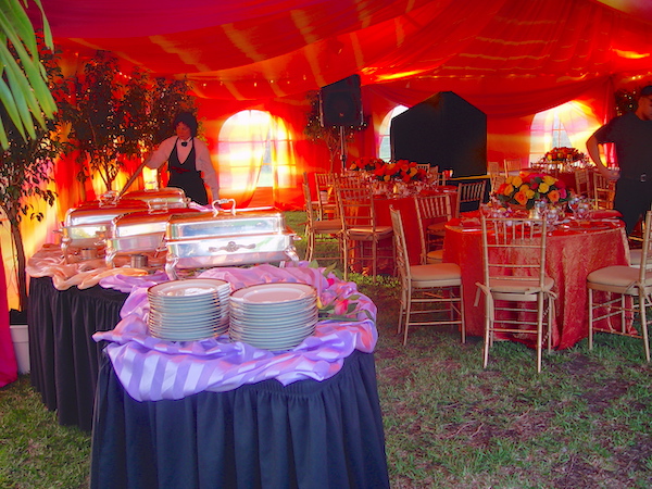 An orange cloth draped tent and dining tables. A curvy purple and lavender draped buffet table set with plates and silver roll top chafing dishes.
