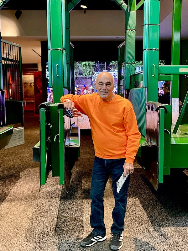 A man in an orange sweater has his arm on the green starting gate at Churchill Downs Louisville Kentucky