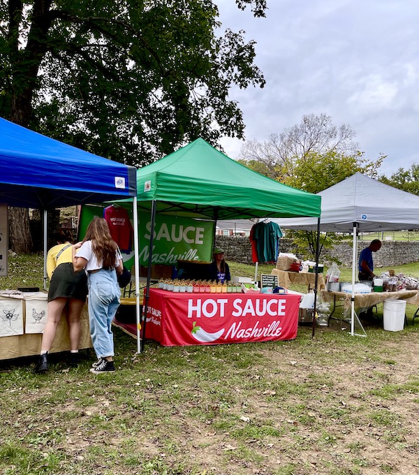 A booth with table draped in red under a green tent. Displayed were bottles of hot sauces at the Farmers Market Nashville TN