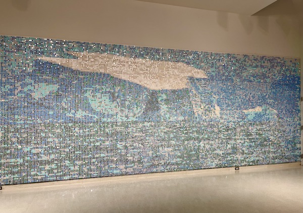 An art installation made of sequins, pins and rubber stoppers depicting sky and clouds takes up an entire wall.