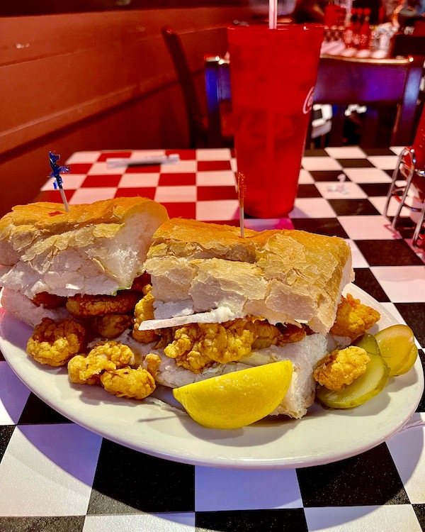 A white platter topped with a fried oyster sandwich with a lemon wedge, sitting on a black and white checkered tablecloth.
