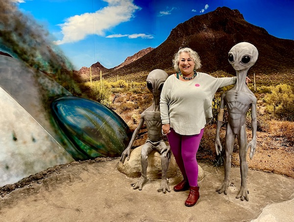 A woman in an ice blue top and magenta leggings stands between 2 alien figures at the International UFO Museum Roswell NM