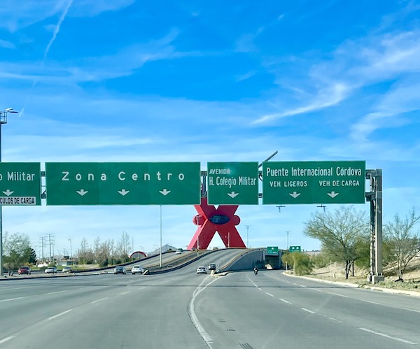 A highway with Mexican exit sights and a bright red cross sculpture from Our Trip to Mexico and Lunch