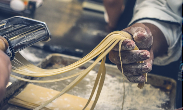 A pasta roller and pasta strands being pulled by a man's hand from The Essentials Behind Italian Cooking
