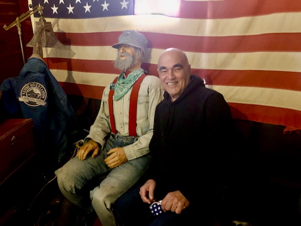 A man in a black sweatshirt sitting next to a mannequin of a train engineer with an American flag behind them at the Verde Canyon Railroad Wilderness Route