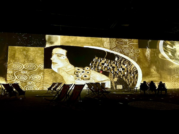 A woman with black hair and a black mole by her eye in gold, cream and black at the Gustav Klimt: The Immersive Experience
