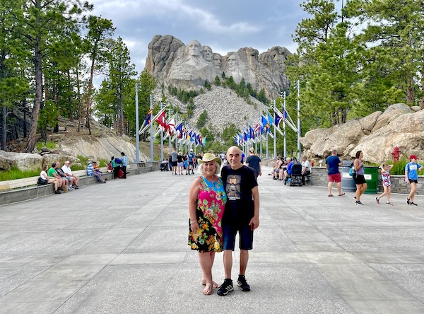 A man in shorts and a woman in a hat and dress in front of Mt. Rushmore wishing you Happy Independence Day 2022!!!