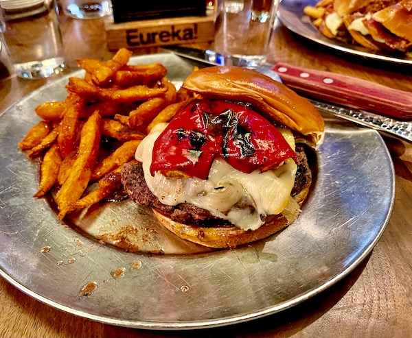 A burger and sweet potato fries on a metal plate. Burger is topped with charred red pepper and melted cheese.