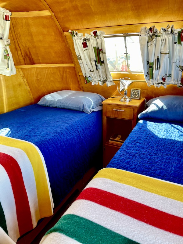 A camper bedroom with twin beds that have red, white, blue and yellow covers at the Palm Springs CA Vintage Trailer Show