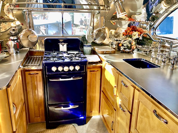 A camper kitchen with light wood cabinets and cobalt blue vintage stove and lots of pots and pans.
