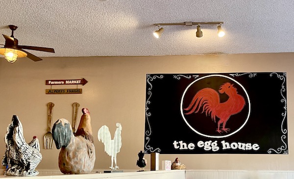 A light beige wall with a sign for the egg house and ceramic roosters at The Egg House Simi Valley CA