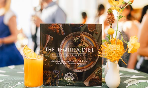 A cookbook flanked by a white vase with yellow flowers and tiny pineapple and an orange colored hi ball with people in the background. The new book is The Tequila Diet by Chef Dave Martin
