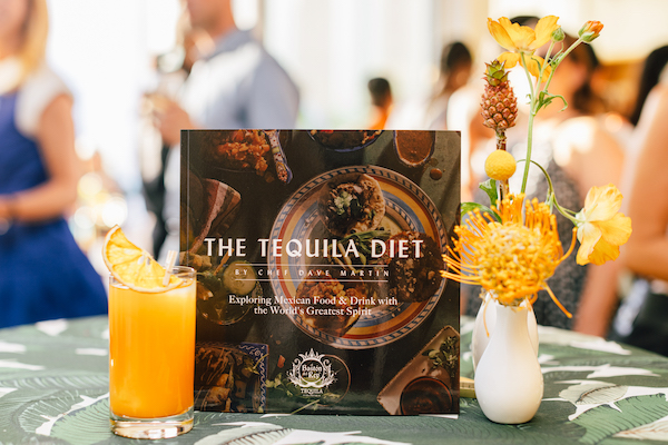 A cookbook flanked by a white vase with yellow flowers and tiny pineapple and an orange colored hi ball with people in the background. The new book is The Tequila Diet by Chef Dave Martin
