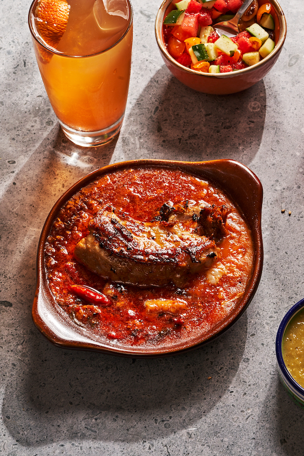 A brown round baking dish filled with red sauce and pork belly. Behind it are a glass of tea colored liquid and a bowl of salad from The Tequila Diet by Chef Dave Martin