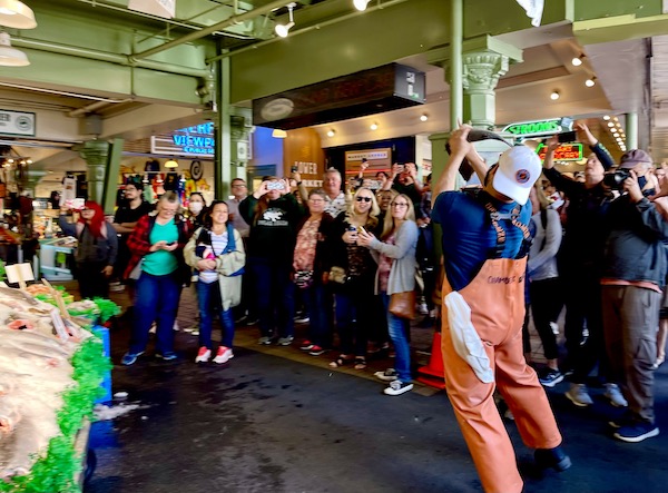 A man in orange coveralls catching a fish that flew through the air at Pike Place Market Seattle WA