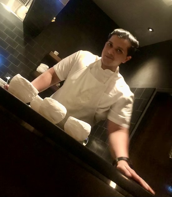 Interview With Chef Karan Puthran in chef whites in a commercial kitchen standing behind a table with a baking sheet and white items on it.