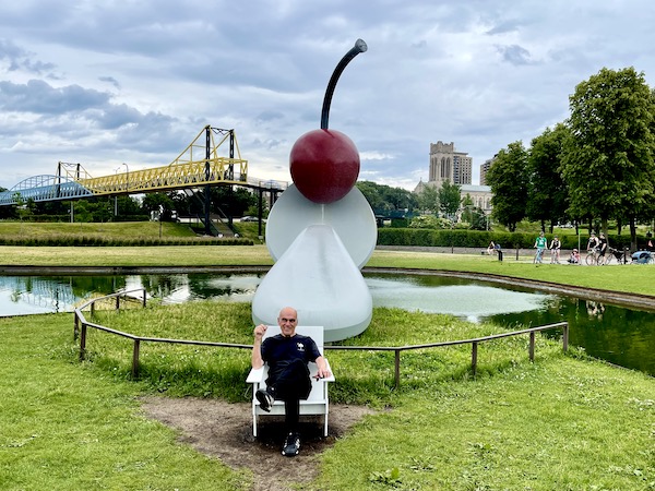 Man in black jeans and t-shirt relaxing on a white Adirondack chair in front of Spoonbridge and Cherry sculpture at Minneapolis Sculpture Garden Minnesota