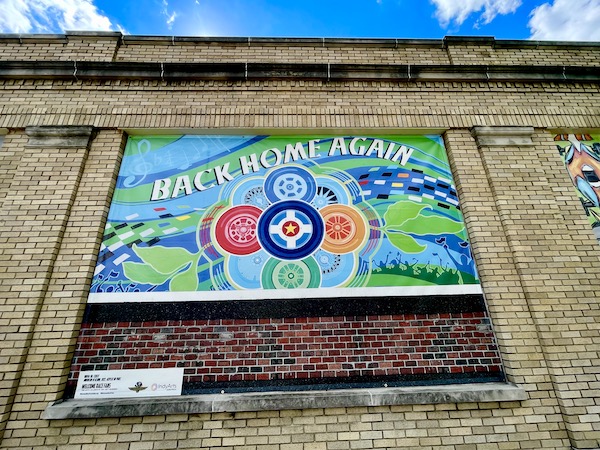 Green, blue and orange mural with wheels welcoming us back to Back Home Again mural at Indianapolis Speedway Indiana with Indy cars.