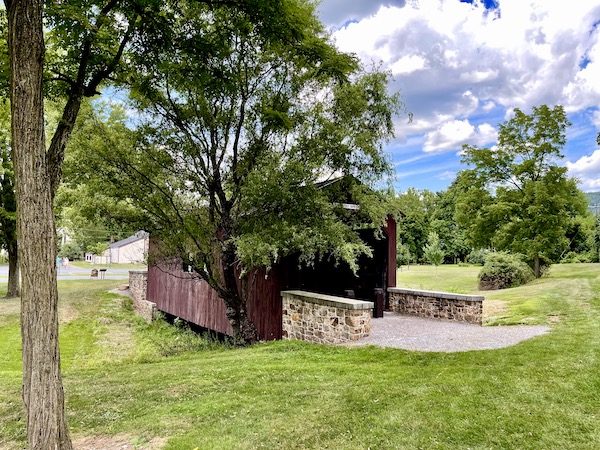 Dark reddish brown wood covered bridge amid trees and lawn.