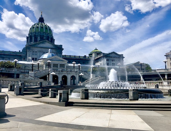 Pennsylvania State Capitol Harrisburg is an oof-white building with columns and green dome. Circular fountains is in front of the steps to the entrance.