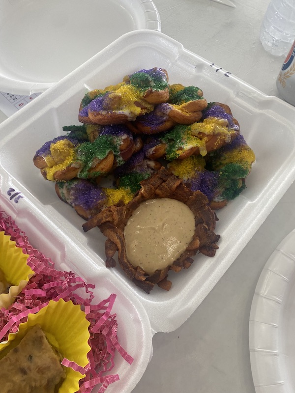 Styrofoam clamshell holding purple green and gold king cake twisted donuts and a bacon bowl filled with sauce.