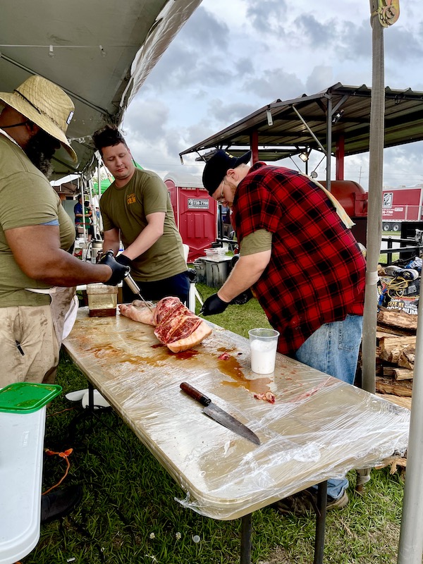 Three men tending to a raw leg of pig and injecting marinade into it at Hogs For The Cause NOLA