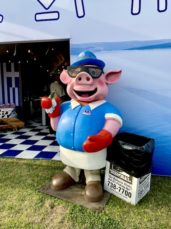 A pig statue with blue cap, short apron and holding a beer.