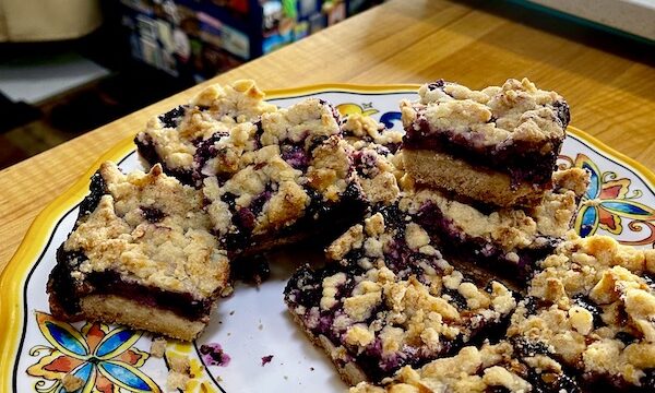Plate of Luscious Blueberry Lemon Bars on a plate sitting on a wood cutting board. Cobalt blue tool chest covered with magnets in the background.