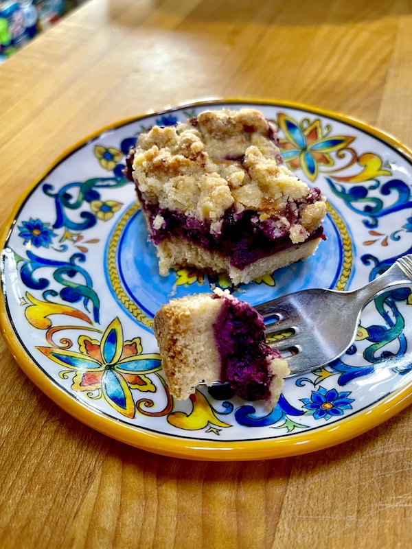 A Luscious Blueberry Lemon Bar on a plate with a forkful taken out