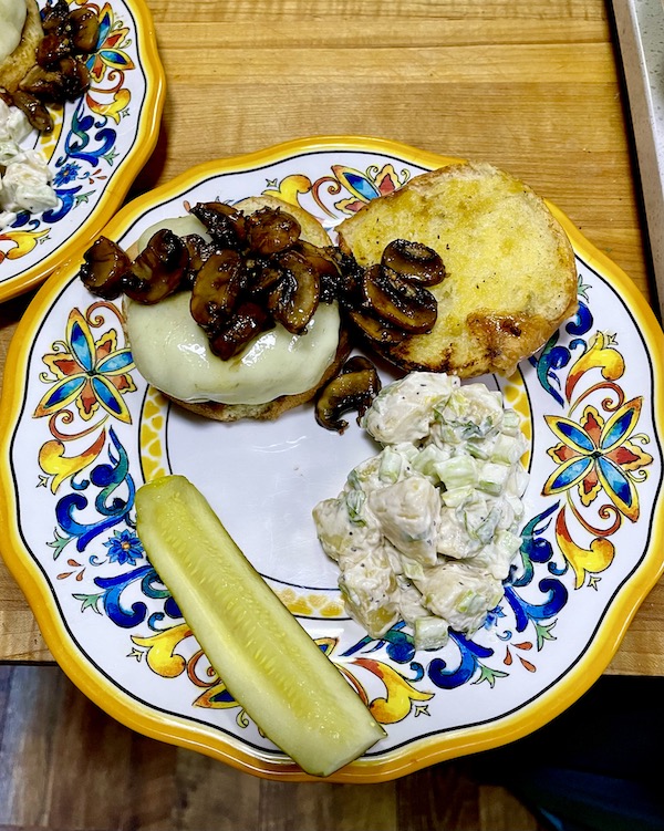 Two colorful plates topped with Mushroom Cheeseburgers, potato salad and a pickle spear