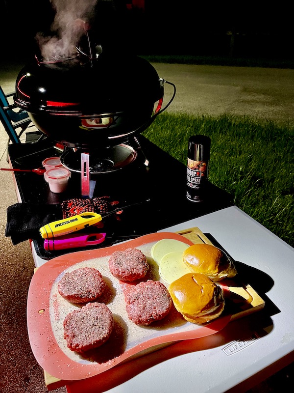 Four raw burger patties, buns and cheese sitting next to the Weber Jumbo Joe Charcoal Grill