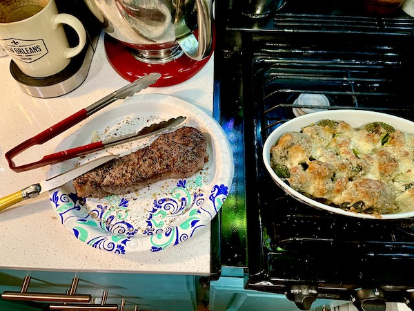 Grilled pork tenderloin on a paper plate with tongs and an oval baking dish filled with baked broccoli cheese casserole sitting on the stove top.