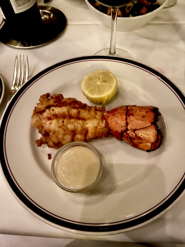 Black rimmed white plate topped with a fried lobster tail, lemon half and small dish of sauce at Chops Lobster Bar Boca Raton