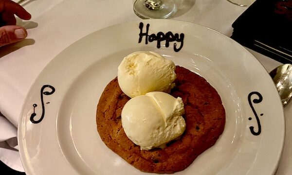 White plate with Happy Birthday written in chocolate on rim and topped with a large chocolate chip cookie and 2 scoops of vanilla ice cream at Chops Lobster Bar Boca Raton