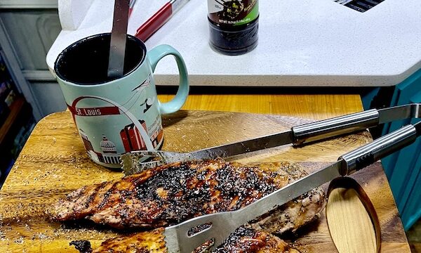 Pair of Easy Glazed Grilled Pork Tenderloin on cutting board with tongs, a mug and jar of sauce on counter.