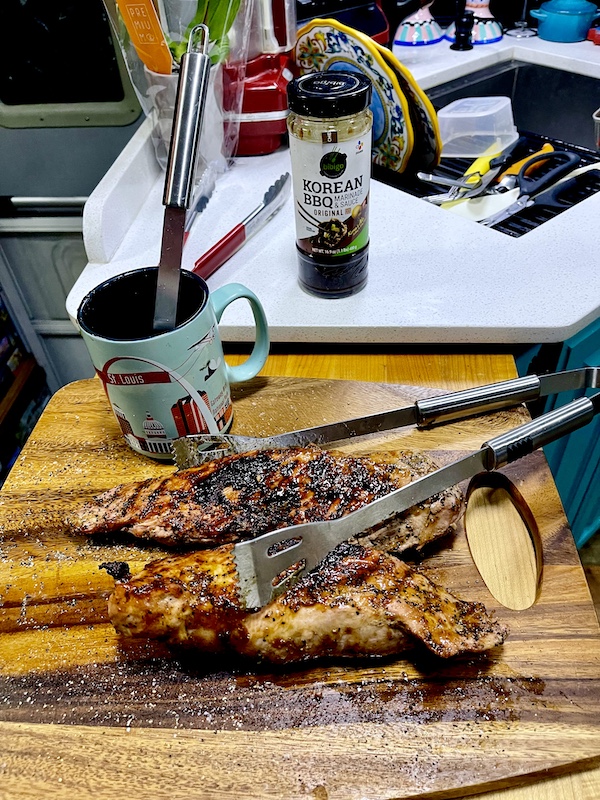 Pair of Easy Glazed Grilled Pork Tenderloin on cutting board with tongs, a mug and jar of sauce on counter.